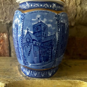 Op de afbeelding: Een blauw-witte keramische pot met een gedetailleerde transferprint van de kathedraal van Carlisle. De pot heeft een cilindrisch lichaam met een deksel, versierd met architecturale illustraties en geometrische patronen, geaccentueerd met goud.