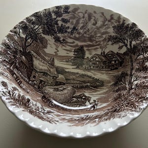 May include: A brown and white ceramic bowl with a rural landscape design. The bowl has a scalloped edge and depicts a village scene with houses, trees, and a horse-drawn cart. The bowl is suitable for serving food.