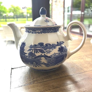 Può includere: Teiera in ceramica bianca con un design paesaggistico blu e bianco. La teiera ha un beccuccio curvo, un corpo arrotondato e un manico ad anello. Il coperchio è sormontato da un pomello blu. Il design presenta ponti, alberi ed edifici.