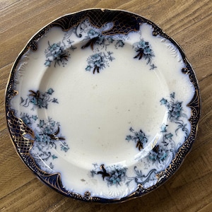 Op de afbeelding: Oude witte bord met een blauw bloemmotief. Het bord heeft een marineblauwe rand met gouden accenten. Het bloemmotief toont blauwe bloemen en bladeren. Het bord is rond en heeft een geschulpte rand.