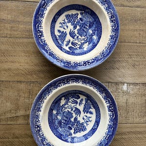May include: Two blue and white ceramic bowls with a classic Willow pattern. The bowls have a wide rim with a detailed border and a central design featuring a landscape scene. The bowls are set on a wooden surface.