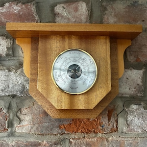 May include: A wall-mounted wooden barometer featuring a circular brass-rimmed gauge with a white face. The dial displays the text Stormy, Rain, Change, Fair, and Very Dry, along with the word Barometer and the brand name Baymaster at the bottom.