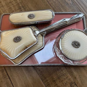 Könnte beinhalten: Ein antikes Kosmetikset mit einem silberfarbenen Tablett und passendem Zubehör. Das Set umfasst einen Handspiegel, eine Bürste und ein rundes Glas, alle mit cremefarbenen Oberflächen und einem zentralen Blumendesign.
