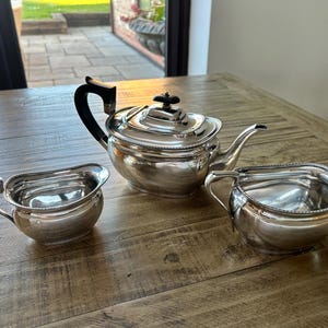 Puede incluir: Un juego de té plateado, que incluye una tetera con asa y tapa negras, una crema y un azucarero. El juego se exhibe sobre una mesa de madera. La tetera tiene forma cuadrada y un pico.