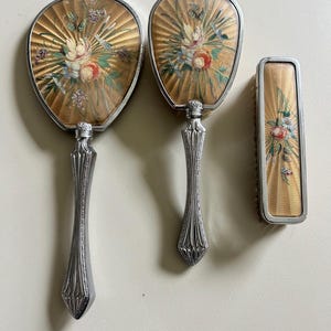 Könnte beinhalten: Ein Vintage-Kosmetikset mit zwei Handspiegeln und einer Bürste. Die Spiegel und die Bürste haben einen silberfarbenen Rahmen und Griff. Die Rückseiten der Spiegel und der Bürste haben einen goldfarbenen Hintergrund mit einem Blumendesign in Weiß, Rot und Blau.