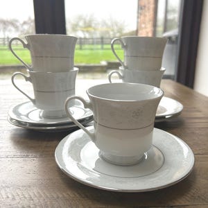 Peut inclure: Un ensemble de tasses et soucoupes en porcelaine blanche. Les tasses et soucoupes présentent un motif floral délicat et un liseré argenté. Les tasses sont empilées, avec un ensemble mis en évidence. L'ensemble est posé sur une table en bois.
