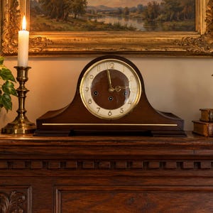 Puede incluir: Un reloj de sobremesa de madera marrón oscuro con parte superior redondeada, esfera circular blanca, números dorados y manecillas doradas. El reloj presenta una línea de incrustación horizontal dorada en la base frontal y dos orificios de cuerda.