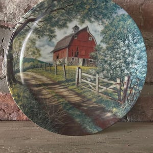 May include: Decorative plate featuring a rural landscape with a red barn, dirt road, and wooden fence. The plate has a glossy finish and is surrounded by a green and blue landscape. The plate is round and has a detailed painting.