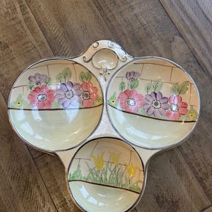 May include: A vintage, three-section serving dish with a cream-colored base and silver trim. Each section features a floral design with pink, purple, and yellow flowers. The dish is likely made of ceramic or porcelain.