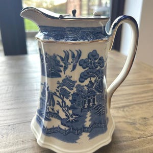 Puede incluir: Jarra de cerámica azul y blanca con tapa, con un diseño de patrón Willow. La jarra tiene un asa curva y ribete dorado. La escena del paisaje es un diseño clásico. Probablemente se utiliza para servir bebidas. Altura aproximada de 20 cm.