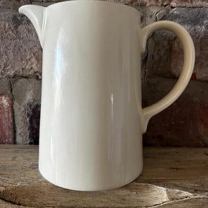 Pode incluir: Um jarro de cerâmica na cor creme com um acabamento liso e brilhante. O recipiente apresenta um corpo cilíndrico, uma alça curva no lado direito e um pequeno bico vertedor integrado na borda superior, oferecendo um design simples e funcional.