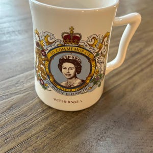 May include: White commemorative mug featuring a portrait of Queen Elizabeth II, with the text "TO COMMEMORATE THE SILVER JUBILEE OF QUEEN ELIZABETH II" and the town name "WITHERNSEA". The mug has a curved handle and a decorative emblem.