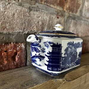 Peut inclure: Une th&eacute;i&egrave;re rectangulaire en c&eacute;ramique orn&eacute;e d'un motif bleu et blanc de type saule. Le d&eacute;cor repr&eacute;sente des pagodes, des ponts et des &eacute;l&eacute;ments paysagers. Elle poss&egrave;de un couvercle assorti avec un bord dor&eacute; et une petite poign&eacute;e sur le dessus.