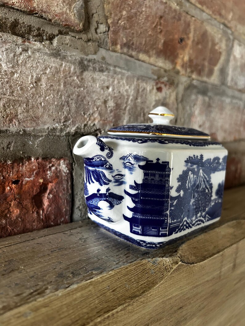 Peut inclure: Une th&eacute;i&egrave;re rectangulaire en c&eacute;ramique orn&eacute;e d'un motif bleu et blanc de type saule. Le d&eacute;cor repr&eacute;sente des pagodes, des ponts et des &eacute;l&eacute;ments paysagers. Elle poss&egrave;de un couvercle assorti avec un bord dor&eacute; et une petite poign&eacute;e sur le dessus.