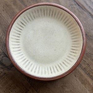 May include: A round ceramic plate with a cream-coloured centre and a brown rim. The plate has a series of vertical grooves radiating from the centre. The plate is sitting on a wooden surface.