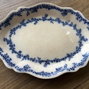 May include: Oval ceramic platter in off-white with a scalloped edge. The rim features a blue floral design. The platter measures approximately 30 cm long and 20 cm wide.