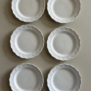 May include: Six white ceramic plates with scalloped edges are arranged in two rows of three. The plates have a simple, elegant design, suitable for serving meals. The plates are clean and appear unused.