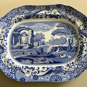 May include: A blue and white ceramic serving platter with an oval shape. The central design depicts a rural landscape with figures and sheep. The border is decorated with floral and scroll patterns. A classic piece.