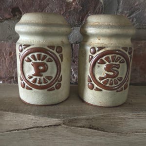 May include: Two ceramic salt and pepper shakers in a beige hue, each featuring a brown circular design. One shaker displays the letter "P", the other "S". The design has a rustic, vintage feel, suitable for a kitchen or dining setting.