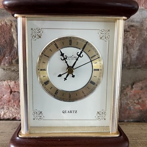 May include: A rectangular quartz clock with a dark brown wooden base and top, and gold-colored metal frame. The clock face is white with gold Roman numerals and hands. The clock has a gold handle at the top.