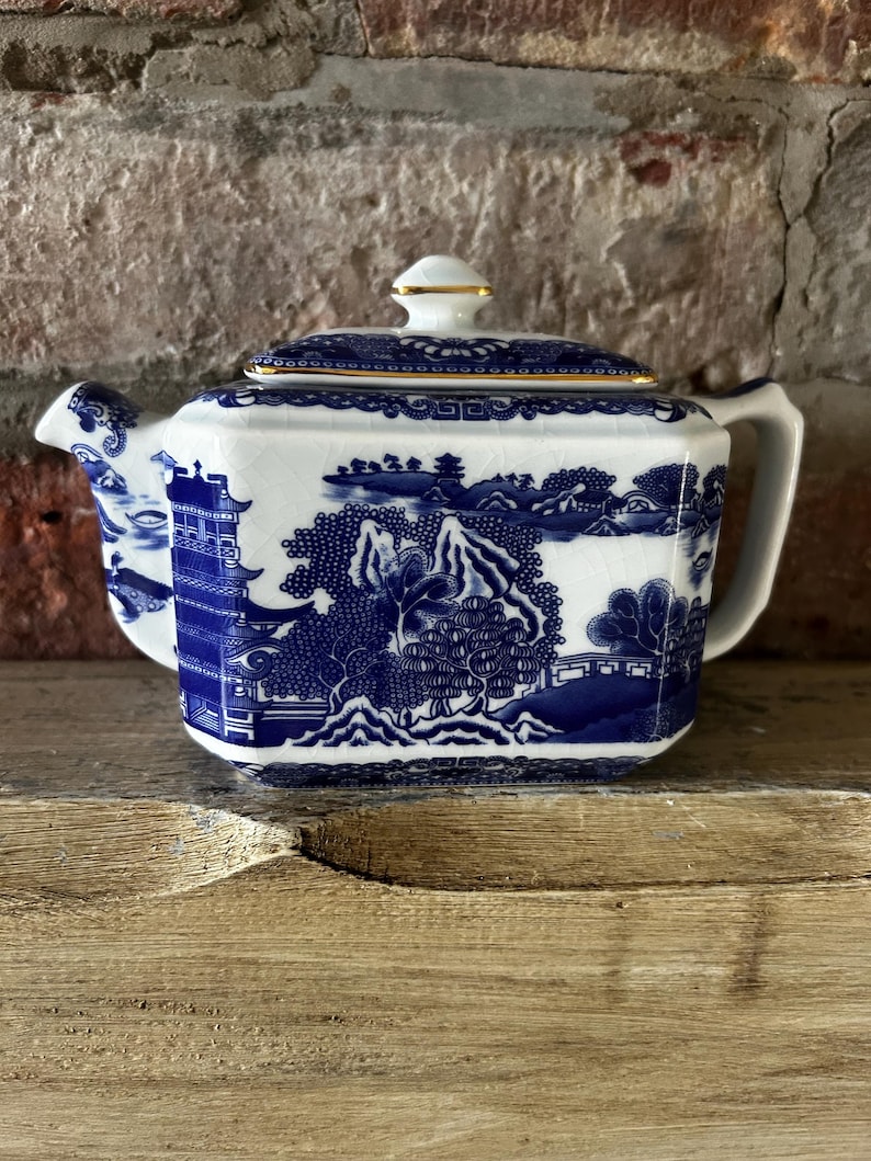 Peut inclure: Une th&eacute;i&egrave;re en c&eacute;ramique blanche avec un motif bleu de type saule repr&eacute;sentant des pagodes, des arbres et des montagnes. La th&eacute;i&egrave;re poss&egrave;de un corps hexagonal, un couvercle assorti avec un bouton dor&eacute;, un bec verseur et une anse.