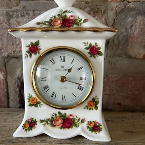 Puede incluir: Un reloj de sobremesa de porcelana blanca con esfera circular de borde dorado, números romanos y el texto Royal Albert Quartz. El reloj presenta un remate decorativo y un patrón floral de rosas rojas y amarillas en el frente.