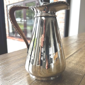 May include: A polished silver-toned pitcher with a tapered body and a wide pouring spout. The handle is wrapped in a light brown woven material. The rim of the pitcher features a decorative beaded edge detail, and the surface is highly reflective.