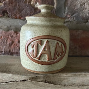 Könnte beinhalten: Ein kleiner, beigefarbener Keramiktopf mit Deckel, mit dem Wort "JAM" in einem braunen Oval. Der Topf hat eine gesprenkelte Textur und steht auf einer Holzoberfläche. Der Hintergrund ist eine Backsteinmauer.