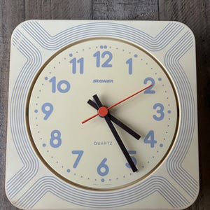 May include: Cream-colored quartz wall clock with blue numerals and hands. The clock face has a cream background with blue numbers and a red second hand. The clock is square with a decorative blue wave pattern around the edge. The clock reads "STAGER QUARTZ".