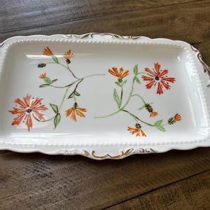 May include: A rectangular white ceramic serving tray featuring a hand-painted orange floral pattern with green stems and leaves. The tray has scalloped edges with a beaded texture and gold-painted accents on the handles and along the rim.