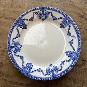 May include: A white ceramic plate featuring a blue floral border design. The plate has a wide rim with a repeating pattern of stylized flowers and scrolls. The plate is round and appears to be vintage or antique.