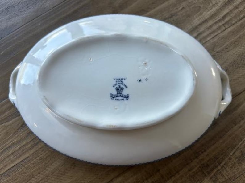Op de afbeelding: Witte ovale serveerschaal met blauwe rand en handvatten. De onderkant heeft een blauw-wit embleem met de tekst "Tuscan Royal Semi Porcelain, England."