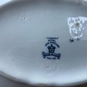 Op de afbeelding: Close-up van een wit porseleinen serveerschaal met een blauw embleem. Het embleem luidt "FORBREX ROYAL SEMI PORCELAIN WOOD & SONS ENGLAND". De schaal is ovaal en vertoont scheuren.