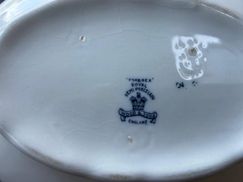 Op de afbeelding: Close-up van een wit porseleinen serveerschaal met een blauw embleem. Het embleem luidt "FORBREX ROYAL SEMI PORCELAIN WOOD & SONS ENGLAND". De schaal is ovaal en vertoont scheuren.