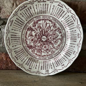 May include: A white decorative plate with a scalloped edge, featuring a calendar for 1982. The plate has a floral design in burgundy and the text "GOD BLESS THIS HOUSE THROUGHOUT 1982".