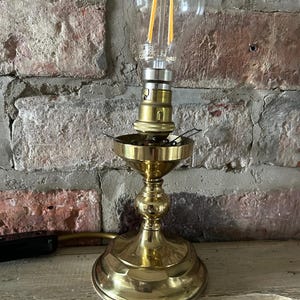 May include: A brass lamp with a clear glass bulb, featuring a decorative base. The bulb has visible filaments. The lamp is set against a brick wall background.