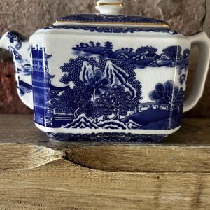 Peut inclure: Une th&eacute;i&egrave;re rectangulaire en c&eacute;ramique &agrave; base blanche orn&eacute;e d'un motif bleu repr&eacute;sentant des pagodes, des arbres et des montagnes. La th&eacute;i&egrave;re poss&egrave;de un couvercle assorti avec un bord dor&eacute; et une finition &eacute;maill&eacute;e craquel&eacute;e sur toute la surface.