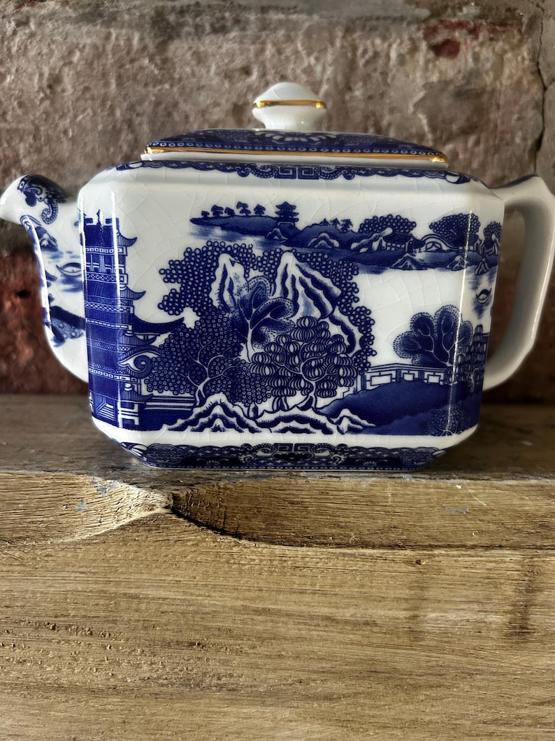 Peut inclure: Une th&eacute;i&egrave;re rectangulaire en c&eacute;ramique &agrave; base blanche orn&eacute;e d'un motif bleu repr&eacute;sentant des pagodes, des arbres et des montagnes. La th&eacute;i&egrave;re poss&egrave;de un couvercle assorti avec un bord dor&eacute; et une finition &eacute;maill&eacute;e craquel&eacute;e sur toute la surface.