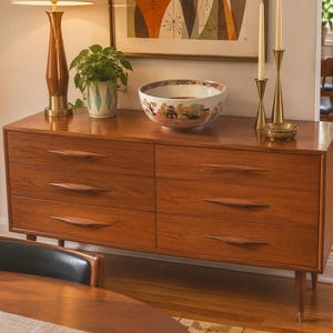 May include: A wooden dresser with six drawers featuring elongated, sculpted handles. On top sits a ceramic bowl with a painted landscape, a potted plant in a patterned pot, a tall lamp with a fabric shade, and two brass candle holders.