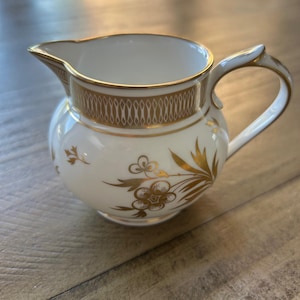 Puede incluir: Una cremera de porcelana blanca con ribetes dorados y diseños florales. La cremera tiene un cuerpo redondeado, un pico y un asa curvada. Los detalles dorados incluyen una banda alrededor de la parte superior y acentos florales.