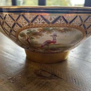 Pode incluir: Uma tigela decorativa de porcelana com borda e base douradas. A tigela apresenta uma pintura detalhada de um pássaro em uma paisagem. O exterior tem um padrão geométrico azul e dourado. Uma peça única para exibição.
