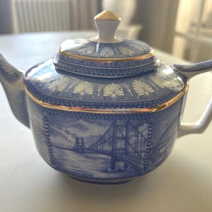 May include: A blue and white ceramic teapot with a lid, handle, and spout. The teapot features a gold trim and a depiction of a bridge on the side. The lid has a gold knob. The teapot is on a white surface.