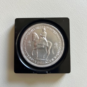 May include: A silver Five Shillings coin, displayed in a black square case. The coin shows a detailed image of a person on horseback, with surrounding text. The coin is a vintage collectible.