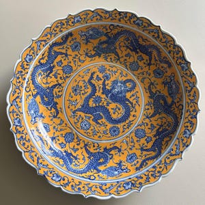 May include: A decorative ceramic plate with a scalloped edge. The plate features a vibrant yellow background with intricate blue dragon and floral designs. The rim is white, and the overall aesthetic is reminiscent of traditional Asian art.