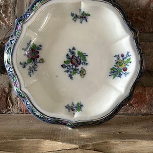 以下が含まれることがあります： 白いセラミック製のサービング皿。縁はスカラップで、花柄のデザイン。皿には、幾何学模様が繰り返される青と金の縁取りがあります。花柄のデザインには、赤、紫、黄色の花が含まれています。
