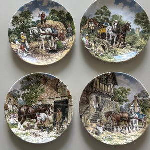 May include: Four decorative plates depicting rural scenes. Each plate shows a different pastoral image of horses, people, and buildings. The plates have a white border and are displayed on a light grey surface.