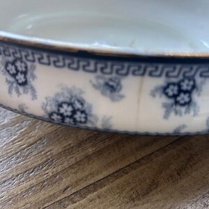 Op de afbeelding: Een witte keramische kom met een blauw bloemmotief en een gouden rand. De kom heeft een herhalend blauw Grieks sleutelontwerp rond de rand. Het bloemmotief omvat clusters van kleine bloemen. De kom staat op een houten oppervlak.