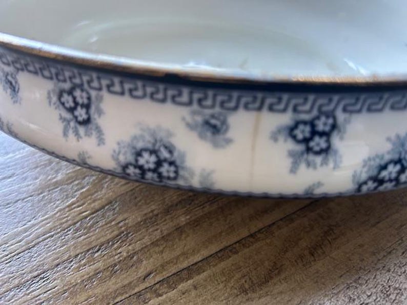 Op de afbeelding: Een witte keramische kom met een blauw bloemmotief en een gouden rand. De kom heeft een herhalend blauw Grieks sleutelontwerp rond de rand. Het bloemmotief omvat clusters van kleine bloemen. De kom staat op een houten oppervlak.