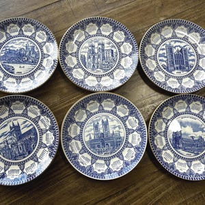 Op de afbeelding: Een set van zes ronde keramische borden met blauw-witte transferware-ontwerpen. Elk bord toont een centrale illustratie van een historische kathedraal of abdij, omringd door een kalender voor een specifiek jaar, waaronder 1956, 1957, 1958, 1959 en 1969.