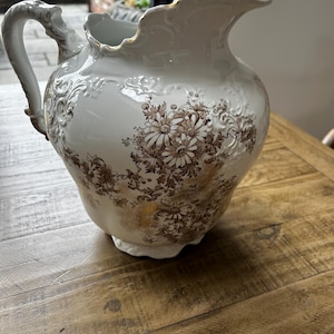 以下が含まれることがあります： 白のセラミックピッチャー。茶色の花柄と金色のアクセントが施されています。ピッチャーは曲線的なハンドルと金色の縁取りが施されたスカラップリムが特徴です。ヴィンテージスタイルの装飾品です。